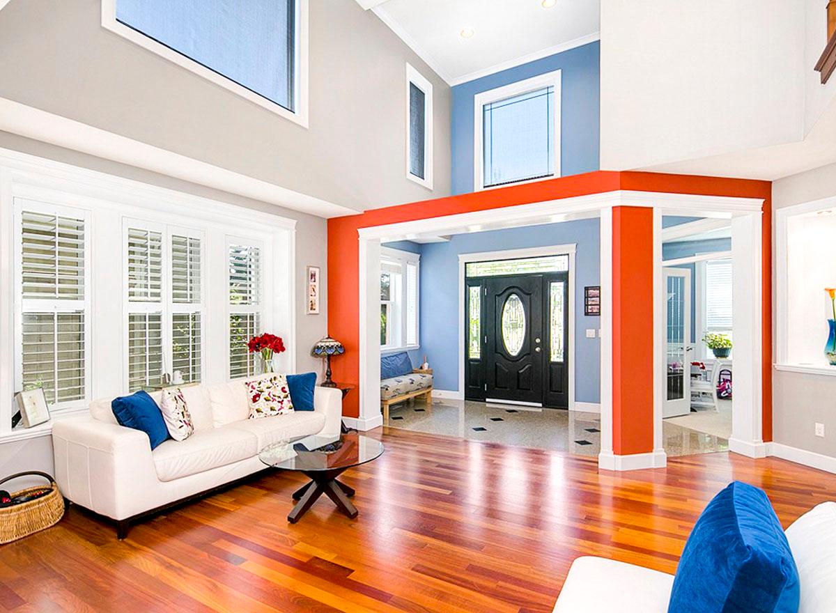 Interior view of a living room and entryway with vaulted ceilings, plantation shutters, a glass coffee table, and a black oval-paneled front door.