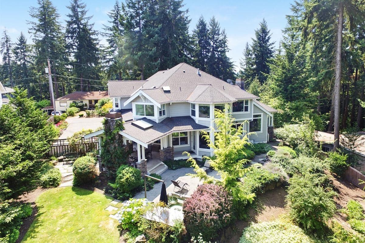 House plan exterior: Two-story home with a covered porch, stone chimney, gables, and bay windows, surrounded by trees.
