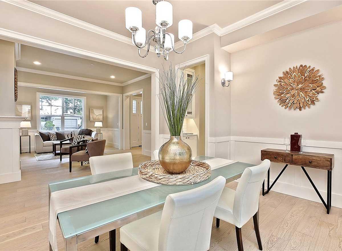 Dining room with glass-top table, white chairs, and adjoining living area. Architectural details include crown molding and wainscoting.