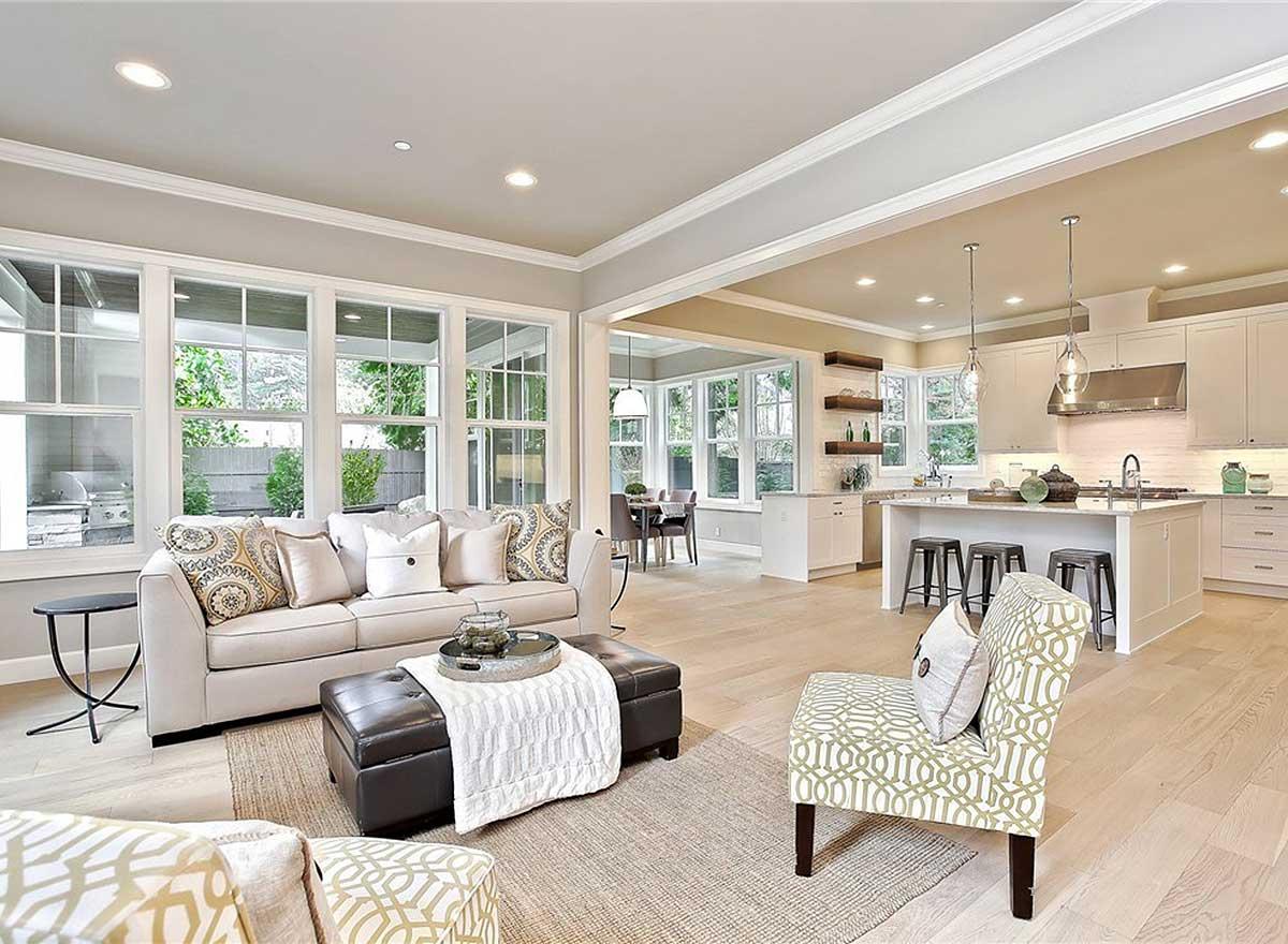 Open-concept living space with large windows, neutral sofa, ottoman, and modern kitchen with island and pendant lights.