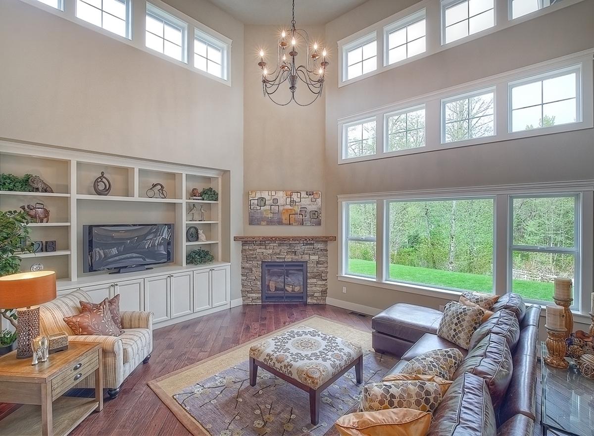 Living room with two-story ceiling, stone fireplace, built-in shelving, and large windows overlooking greenery.
