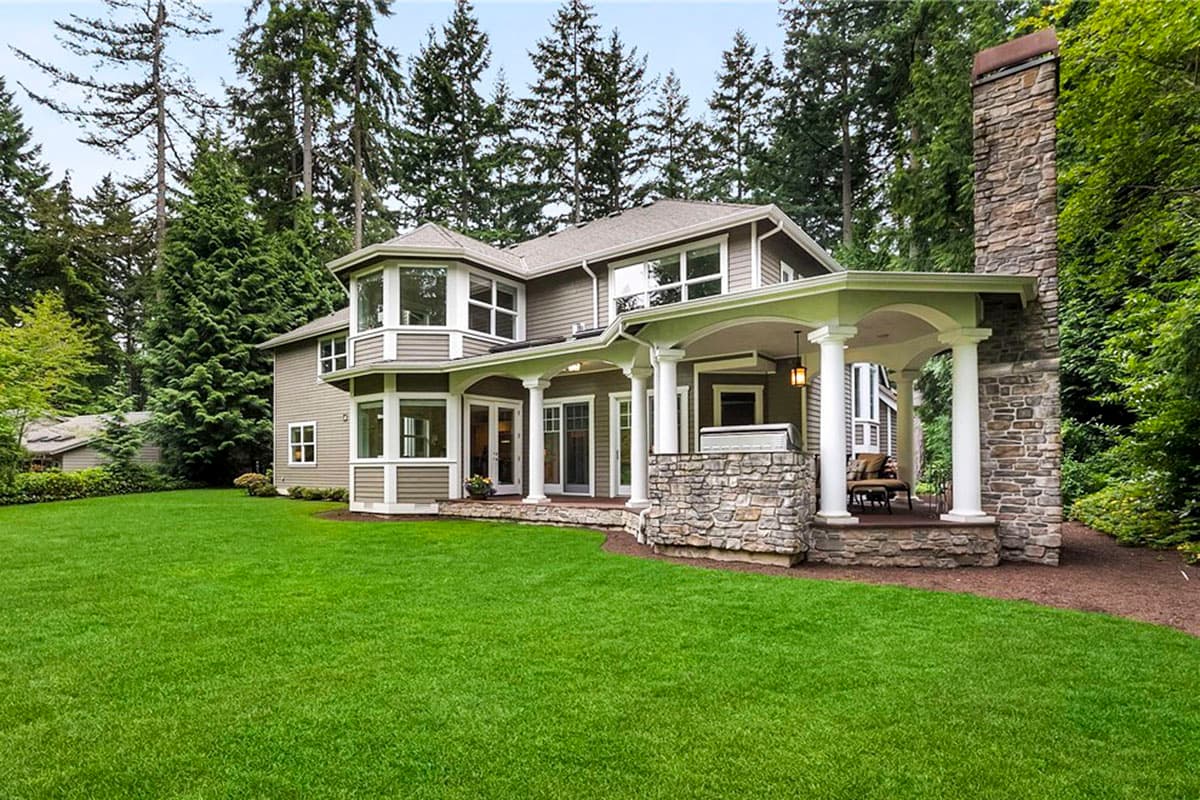 Two-story house plan exterior with covered patio, stone base, bay windows, large chimney, and manicured lawn.