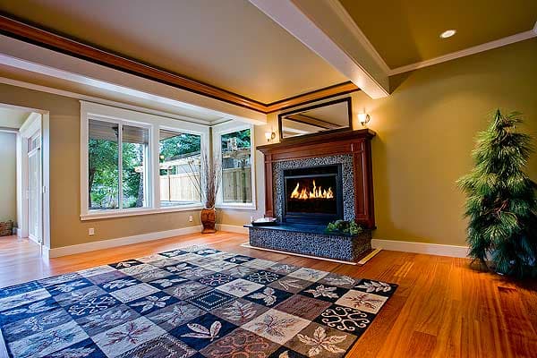 Interior view of living room with fireplace, large windows, and patterned rug on hardwood floor.