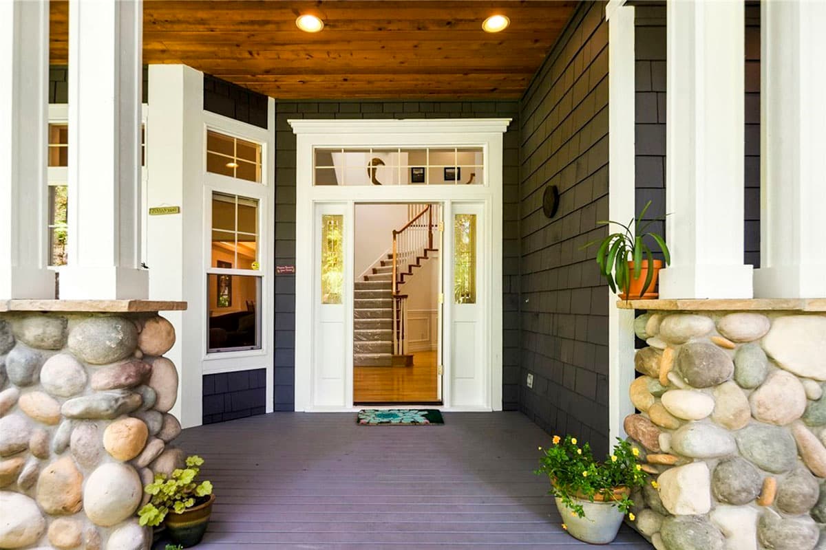 House plan exterior: covered porch with stone-clad columns, dark shakes siding, and double front doors with sidelights leading to stairs.
