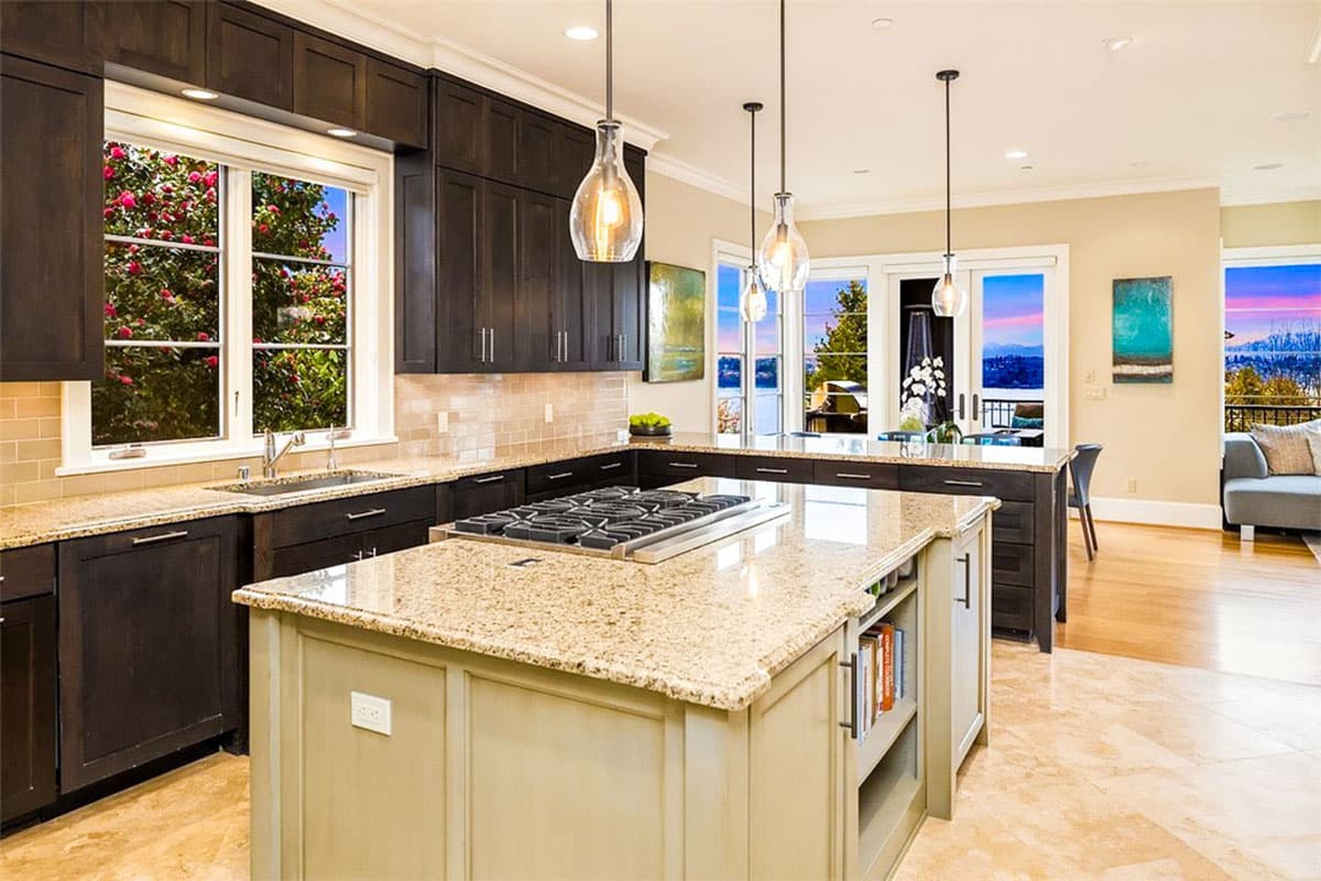 Contemporary kitchen with dark cabinets, granite countertops, large island with gas range, and wide windows overlooking a scenic view.