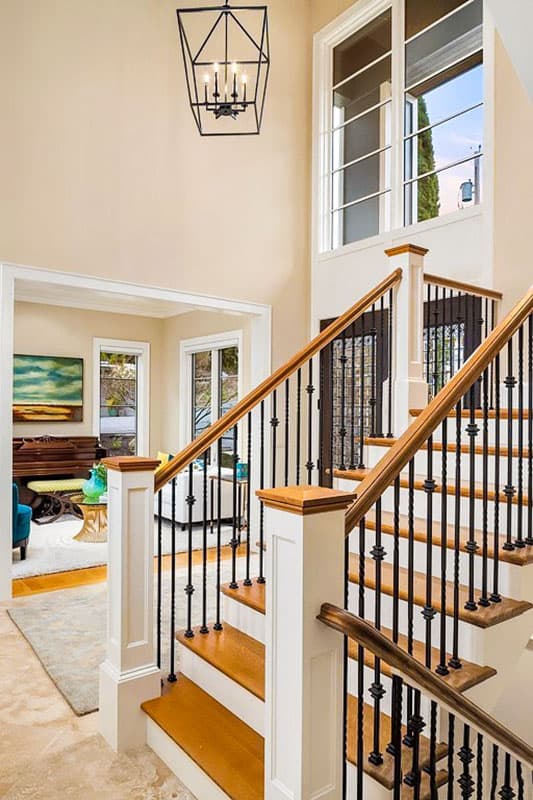 Interior view of a grand staircase with wood treads, black metal balusters, and a tall window overlooking a living area.