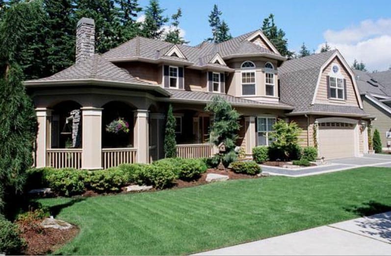 House plan exterior: Two-story home with a wraparound porch, stone chimney, arched dormers, and a side-entry garage.