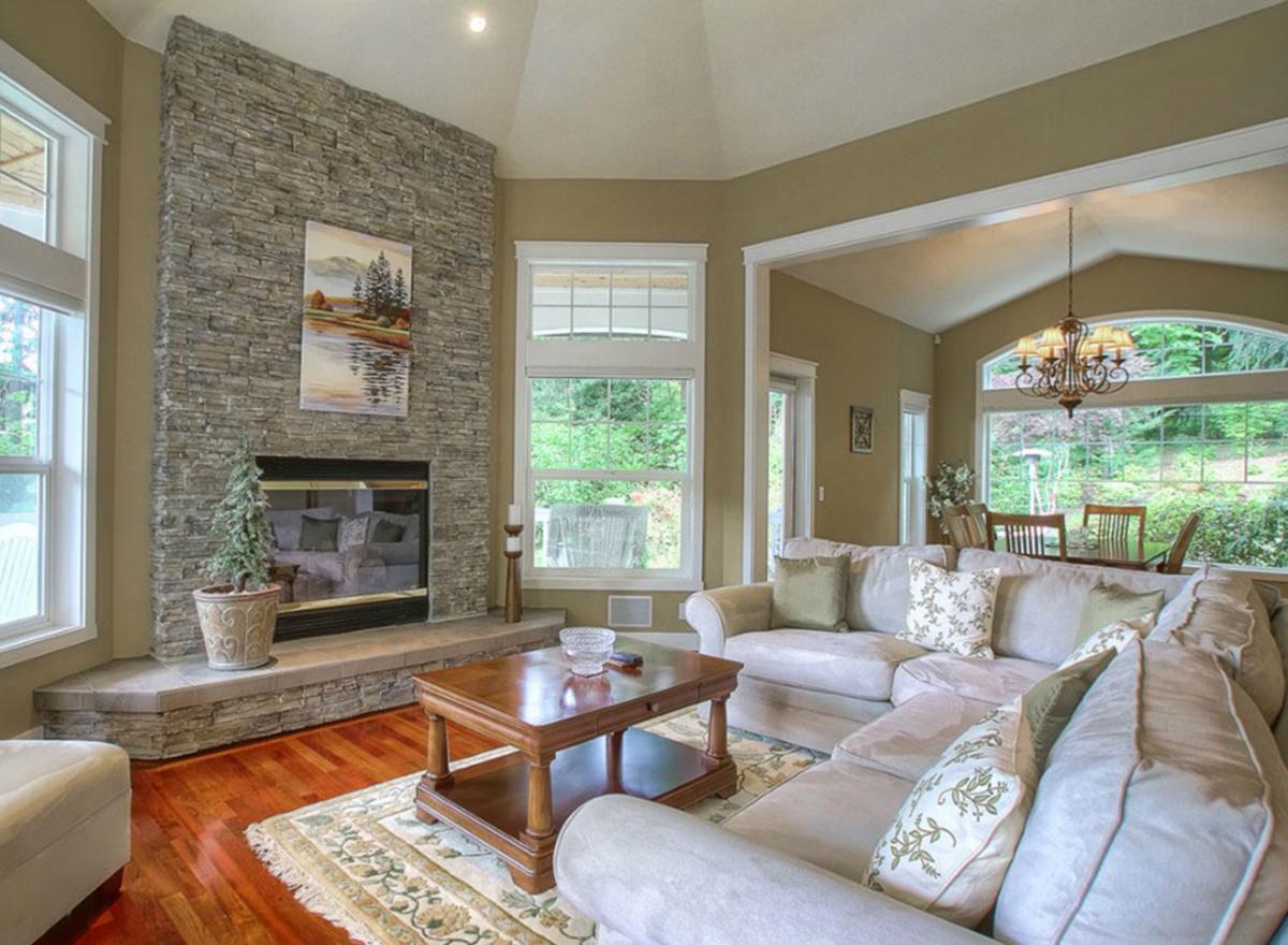 Living room interior with stone fireplace, vaulted ceiling, sectional sofa, coffee table, and large windows overlooking backyard.
