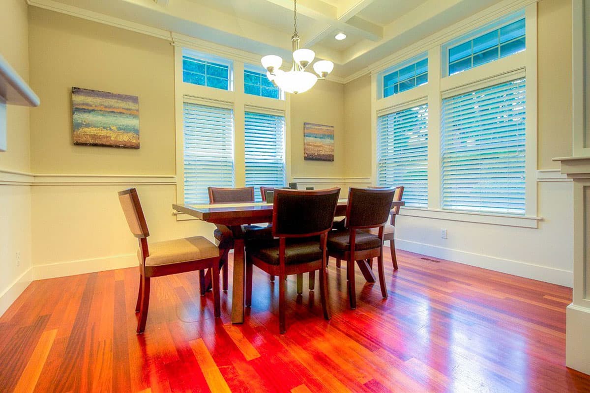Dining room interior with dark wood table and chairs, coffered ceiling, and large windows with blinds.