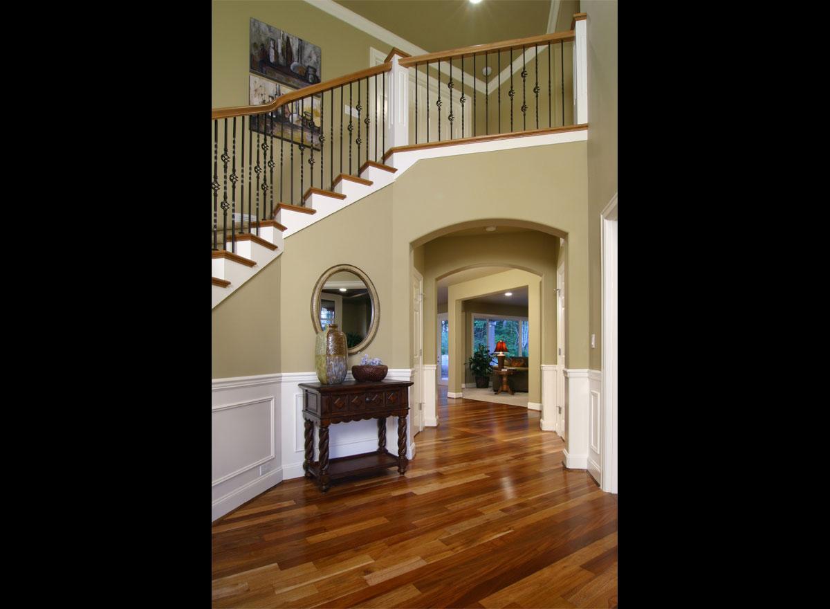 Interior view of a foyer with a staircase, ornate balusters, wainscoting, arched doorways, and hardwood flooring.