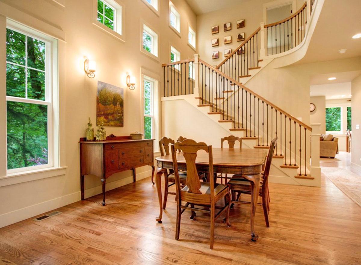 Interior view of dining area with wooden table and chairs, featuring a grand staircase with ornate balusters and a wall of windows.