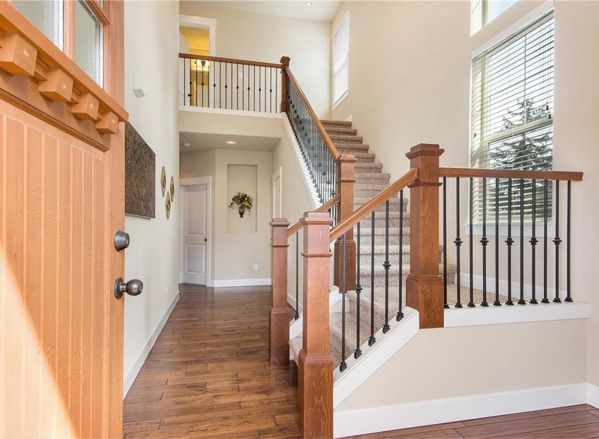 Interior view of house entryway with wood flooring, staircase with wood banisters and metal spindles, and high ceilings.