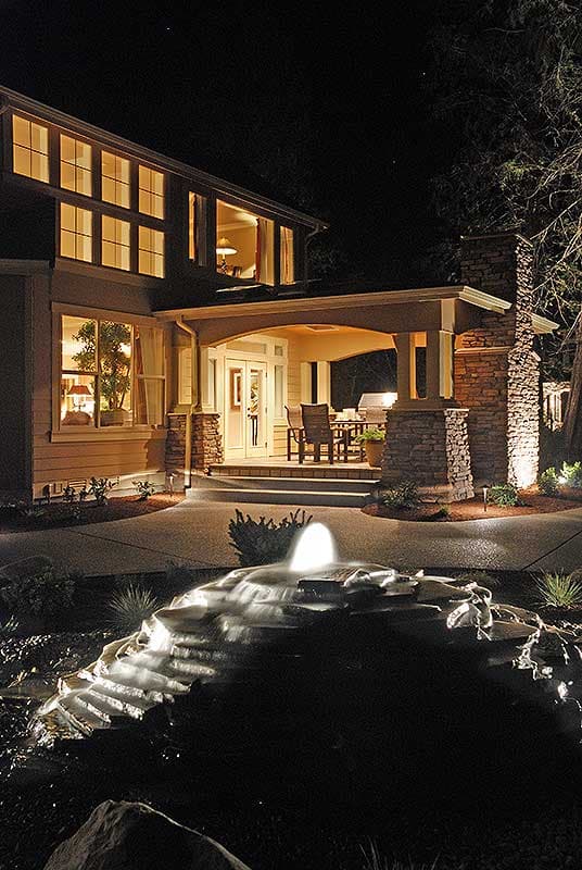 Two-story house plan exterior with covered porch, large stone chimney, and multi-pane windows. Fountain in foreground.
