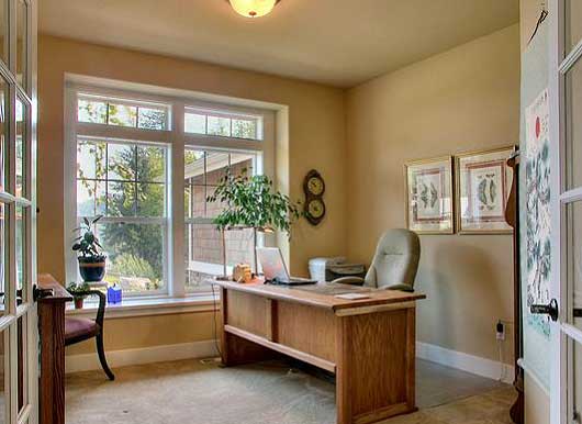 Office room with large windows, wooden desk, chair, and a laptop. Decor includes framed art and a plant.