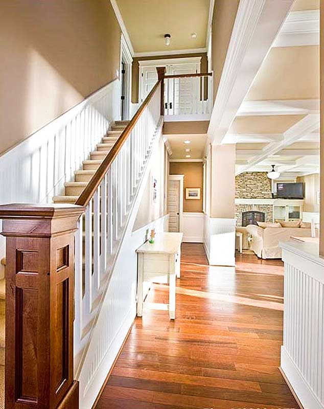 Interior view of a staircase and hallway with wainscoting, hardwood floors, and view into a living room with stone fireplace.