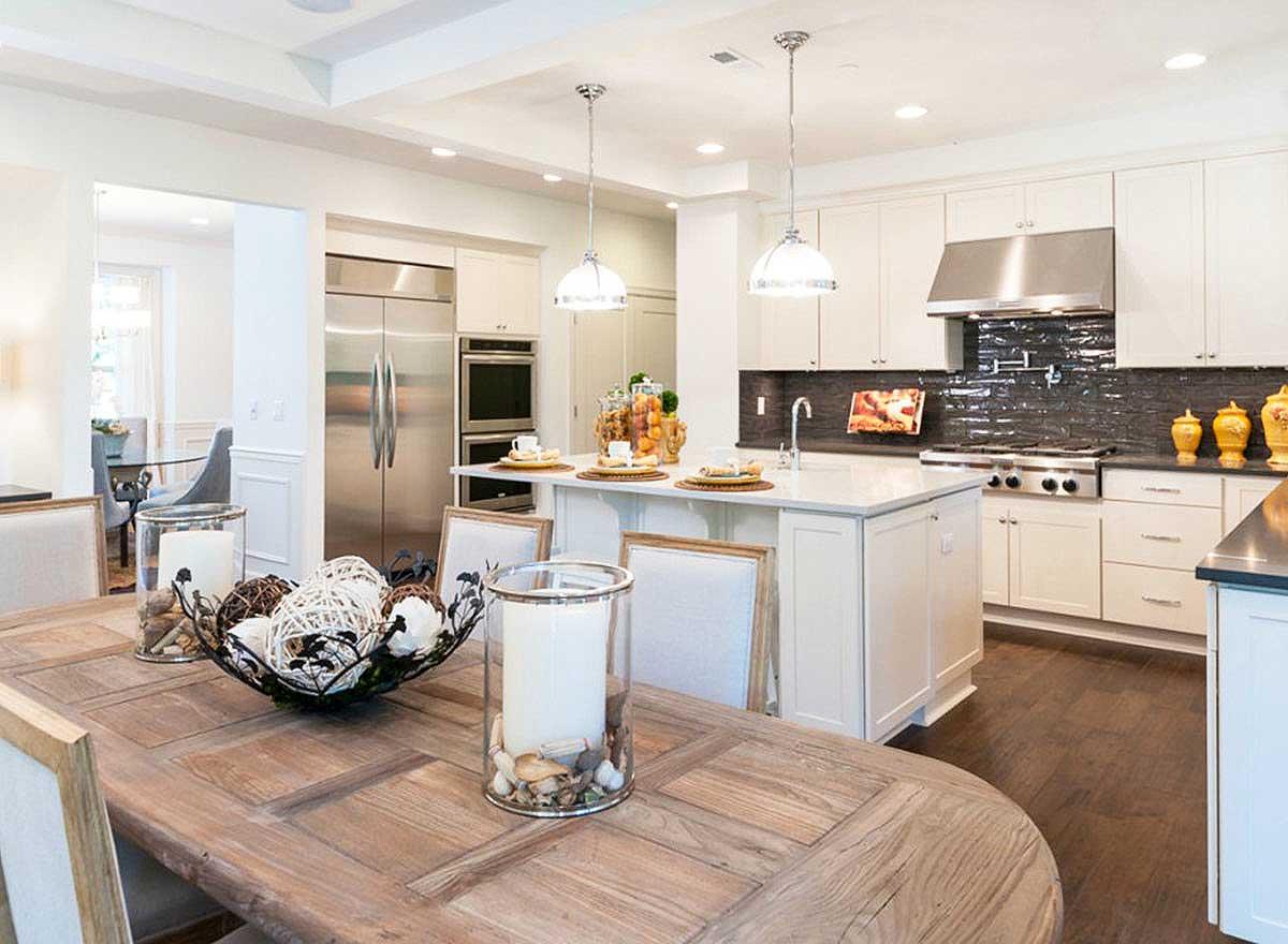 Open concept kitchen with island, stainless steel appliances, white cabinets, dark tiled backsplash, and adjoining dining area.