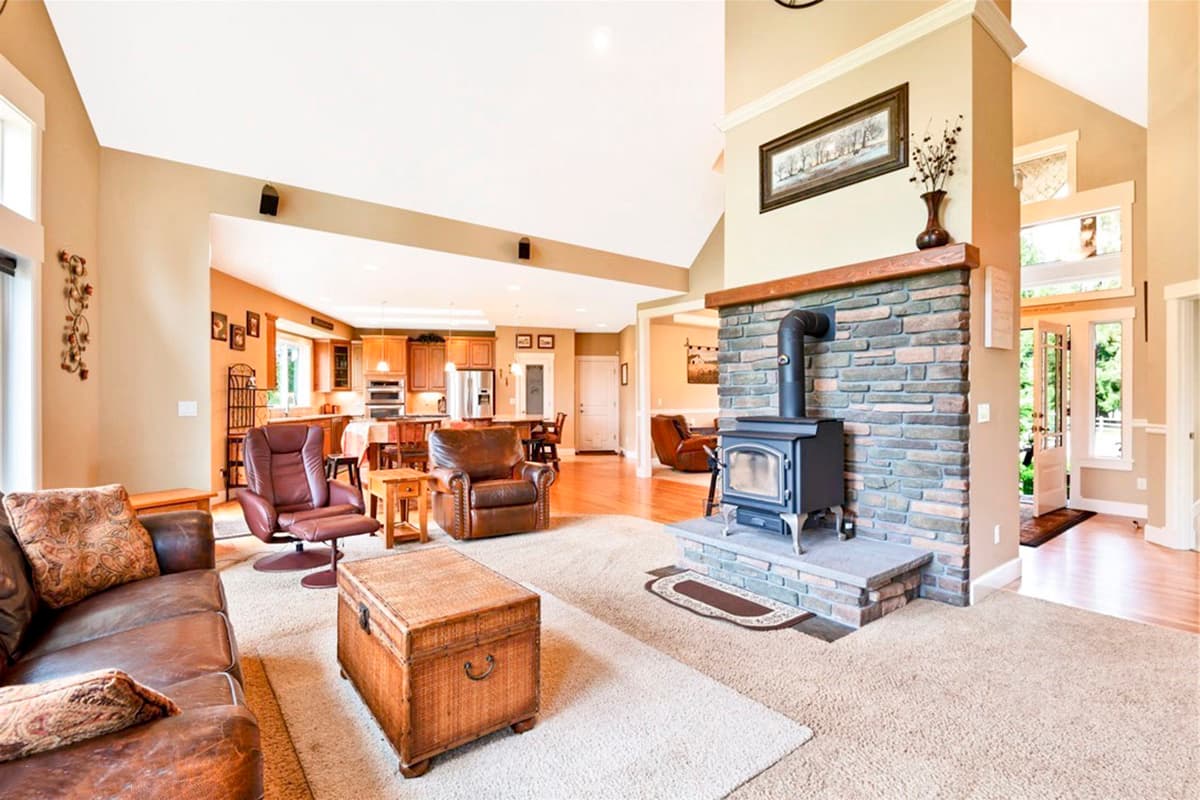 Interior view of a living room with a stone fireplace, wood stove, and open layout to the kitchen and dining area.