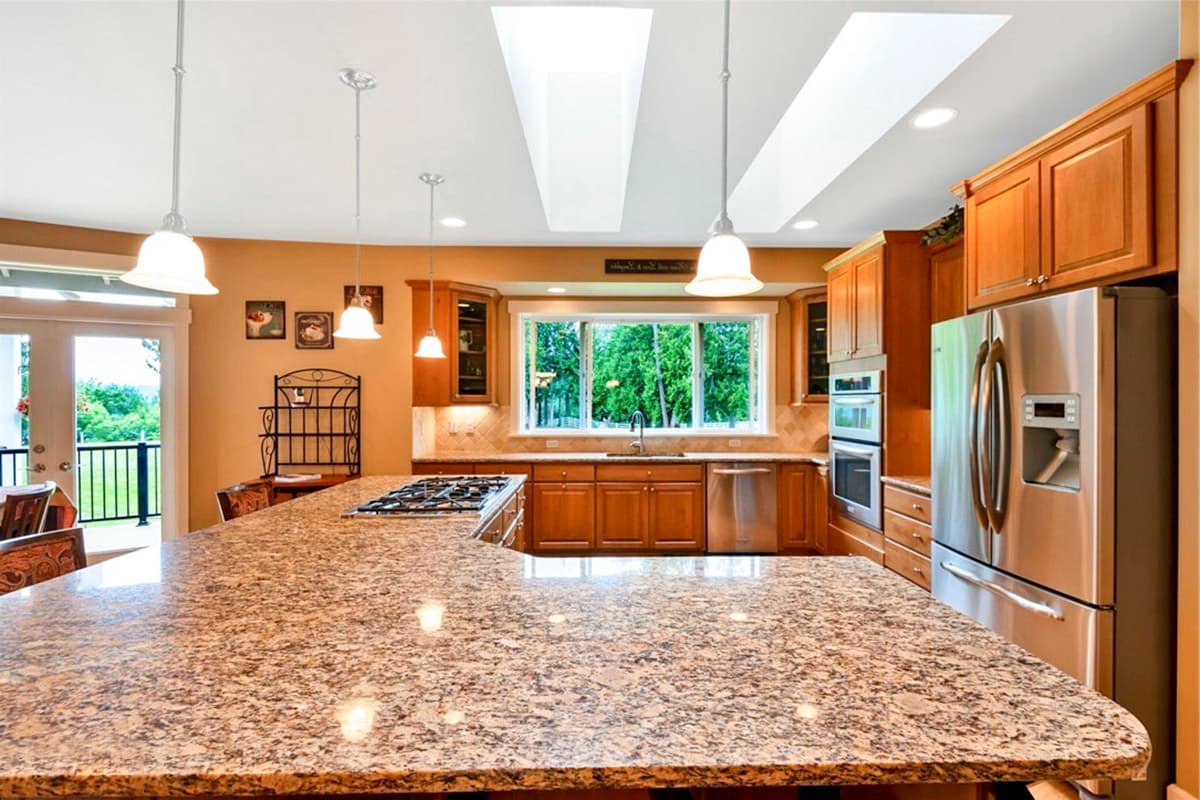 Kitchen with large granite island, stainless steel appliances, wood cabinetry, and a gas range. Skylights illuminate the space.