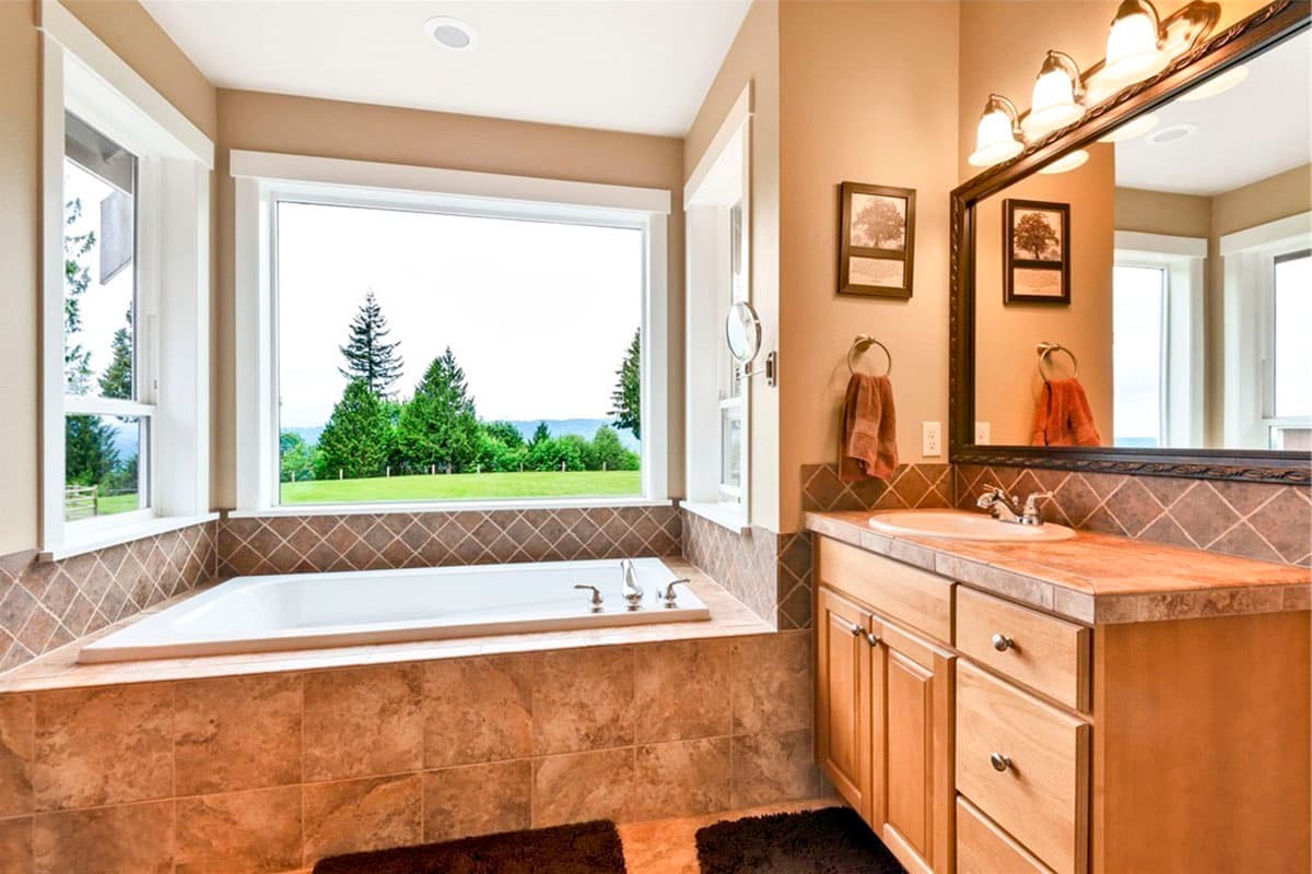 Bathroom with soaking tub and large windows overlooking a green landscape. Double vanity with wooden cabinets is visible.