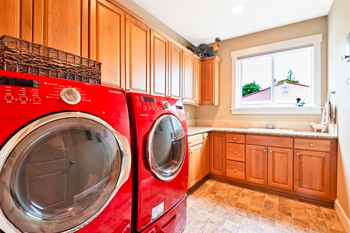 Laundry room with red front-loading washer and dryer, wood cabinetry, granite countertop, sink, and window.