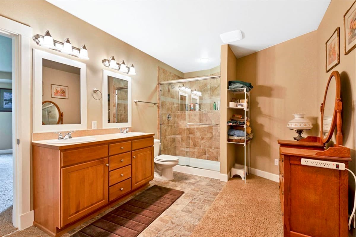 Bathroom interior featuring a double vanity with wood cabinets, glass-enclosed shower with tiled walls, and carpeted flooring.