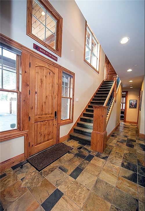Interior view of a spacious entryway with a large wooden double door, slate tile flooring, and carpeted stairs leading to the upper level.