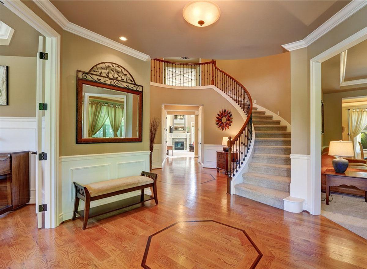 Foyer with curved staircase, wainscoting, and inlaid wood floor. View through doors to living room with fireplace.
