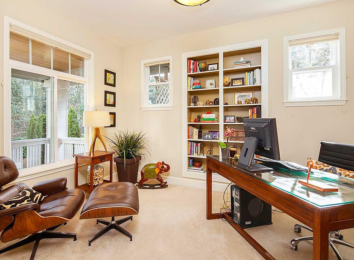 Home office with desk, computer, bookshelves, leather chair, and ottoman. Large windows reveal a porch and trees.