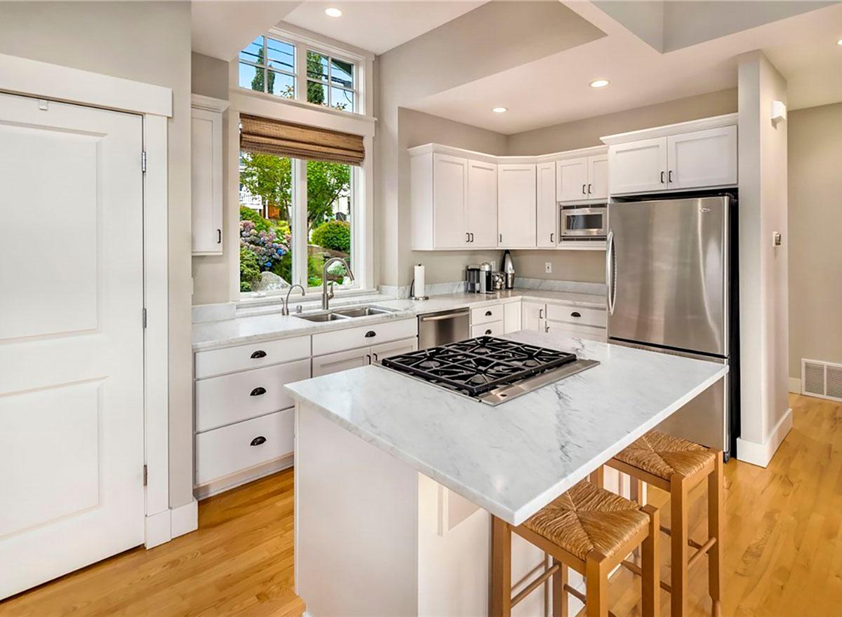 Kitchen with white cabinetry, marble island with gas range, stainless steel appliances, and a large arched window overlooking a garden.