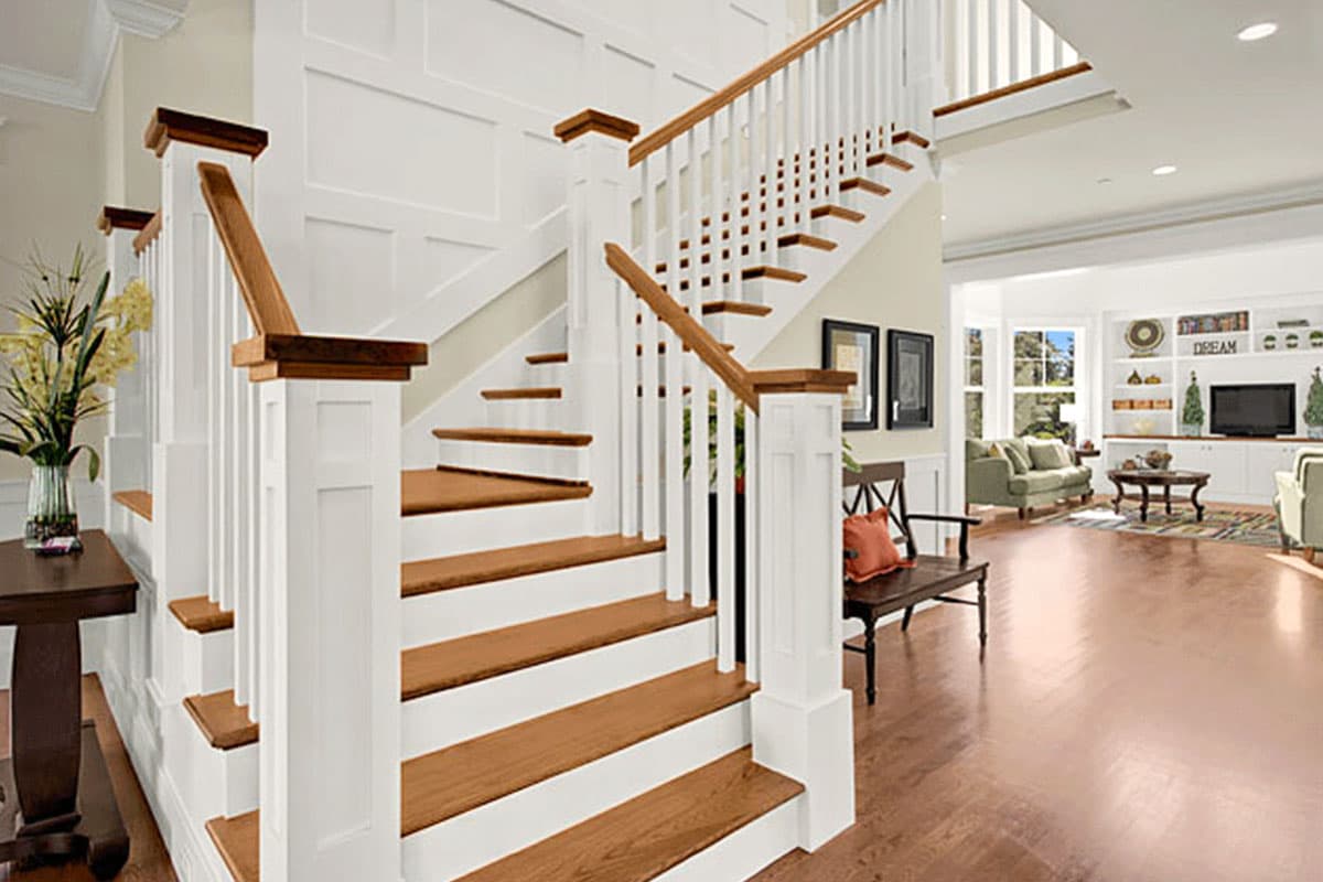 Interior view of a grand staircase with white balusters, wood treads, and paneled wainscoting, leading to a living area.