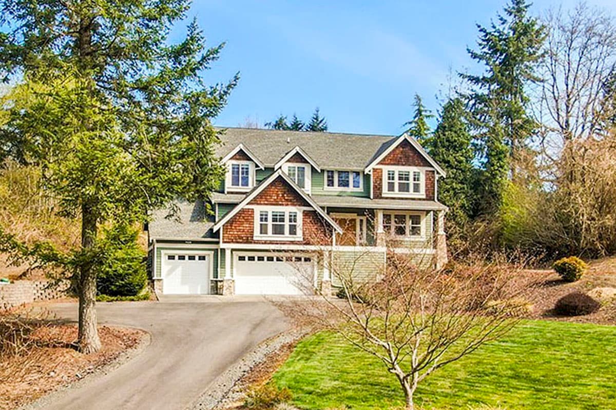 House plan exterior: two-story, green siding, cedar shingles, gables, dormers, covered porch, and a four-car garage.