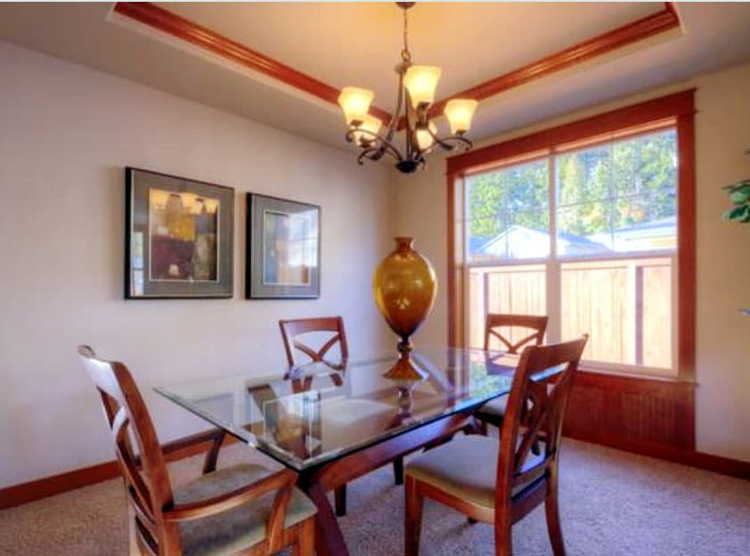 Dining room with glass-top table, wooden chairs, two framed artworks, and tray ceiling with wood trim.