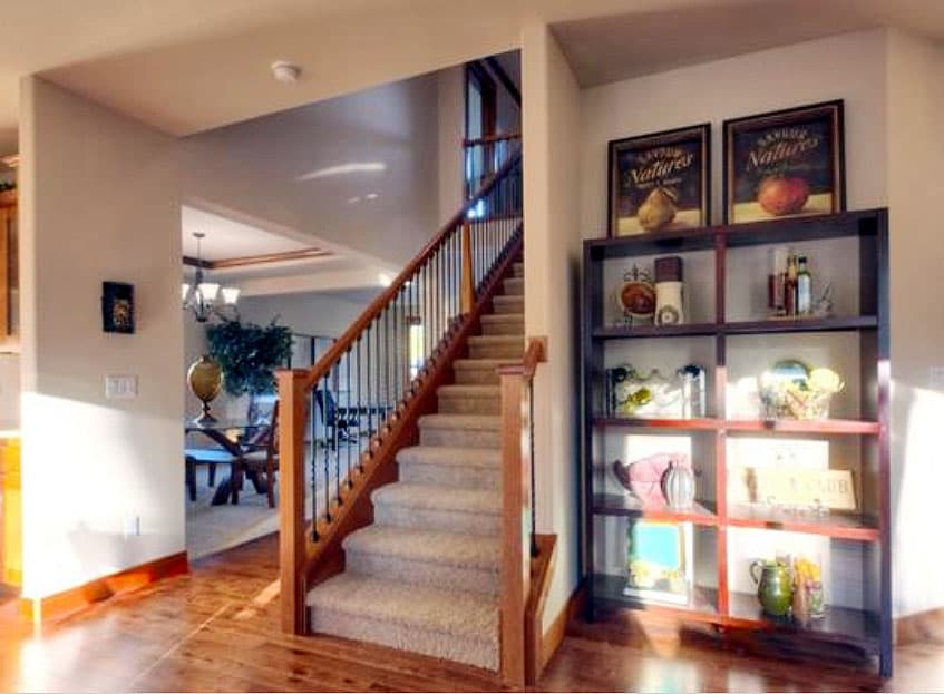 Interior view of house with carpeted staircase, wood banister, and open doorway to dining area. Tall bookshelf visible.