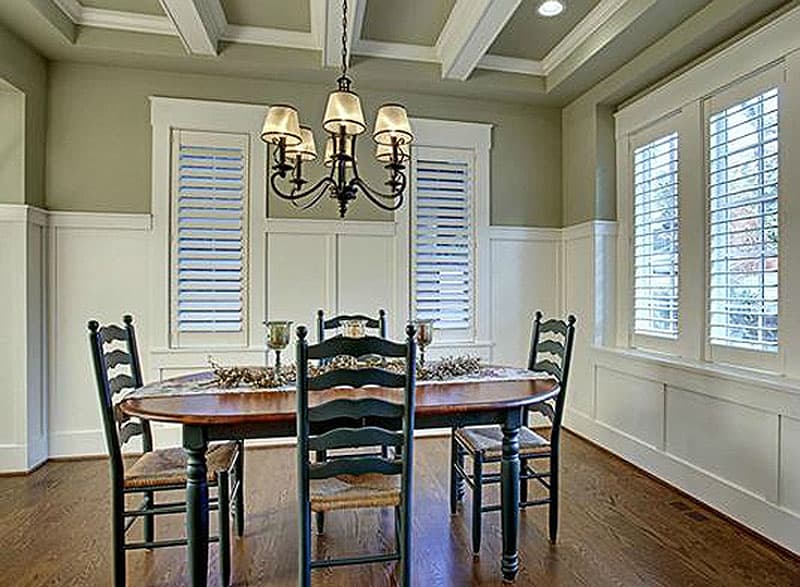 Dining room with coffered ceiling, wainscoting, shuttered windows, and oval dining table.
