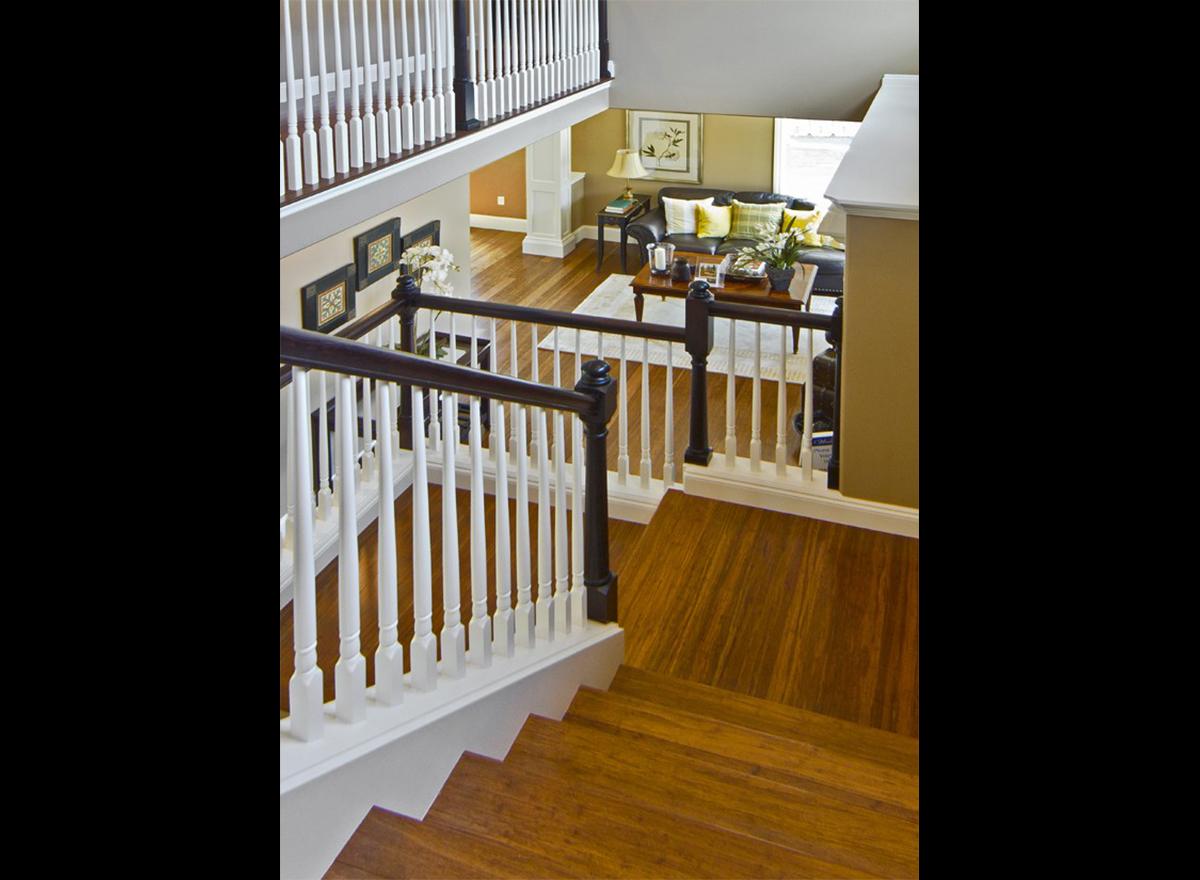 Interior view of a house showing a staircase with white spindles, dark wood railing, and a living room with dark leather sofa and coffee table below.