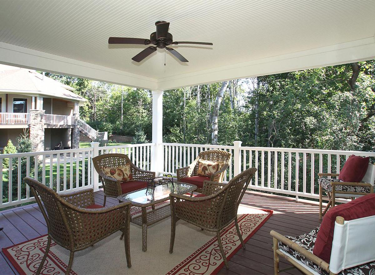 Covered deck with white railing, ceiling fan, and seating, overlooking wooded area and house with stone accents.