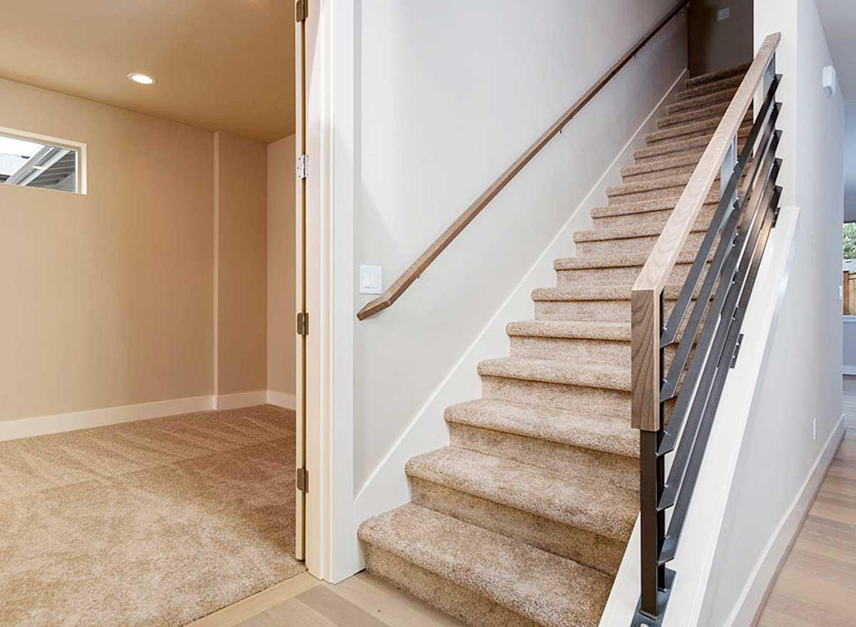 Interior view of a staircase with carpeted treads, wooden handrail, and modern metal balusters. Doorway to carpeted room on left.