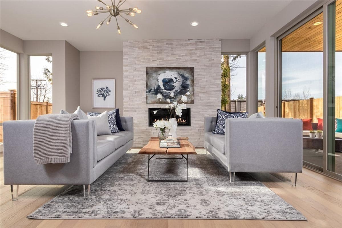 Living room with two gray sofas flanking a stacked stone fireplace, large windows, and sliding glass doors leading outdoors.