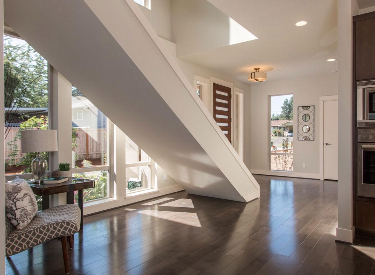 Interior view of modern house with open layout, large windows, and floating staircase. Wood floor reflects sunlight.