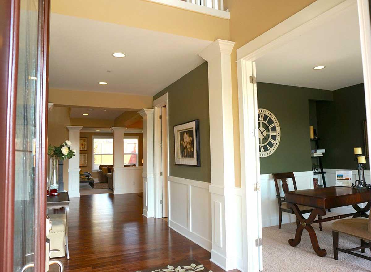 Interior view of a house entryway with hardwood floors, columns, and an open doorway to a study with a desk and clock.