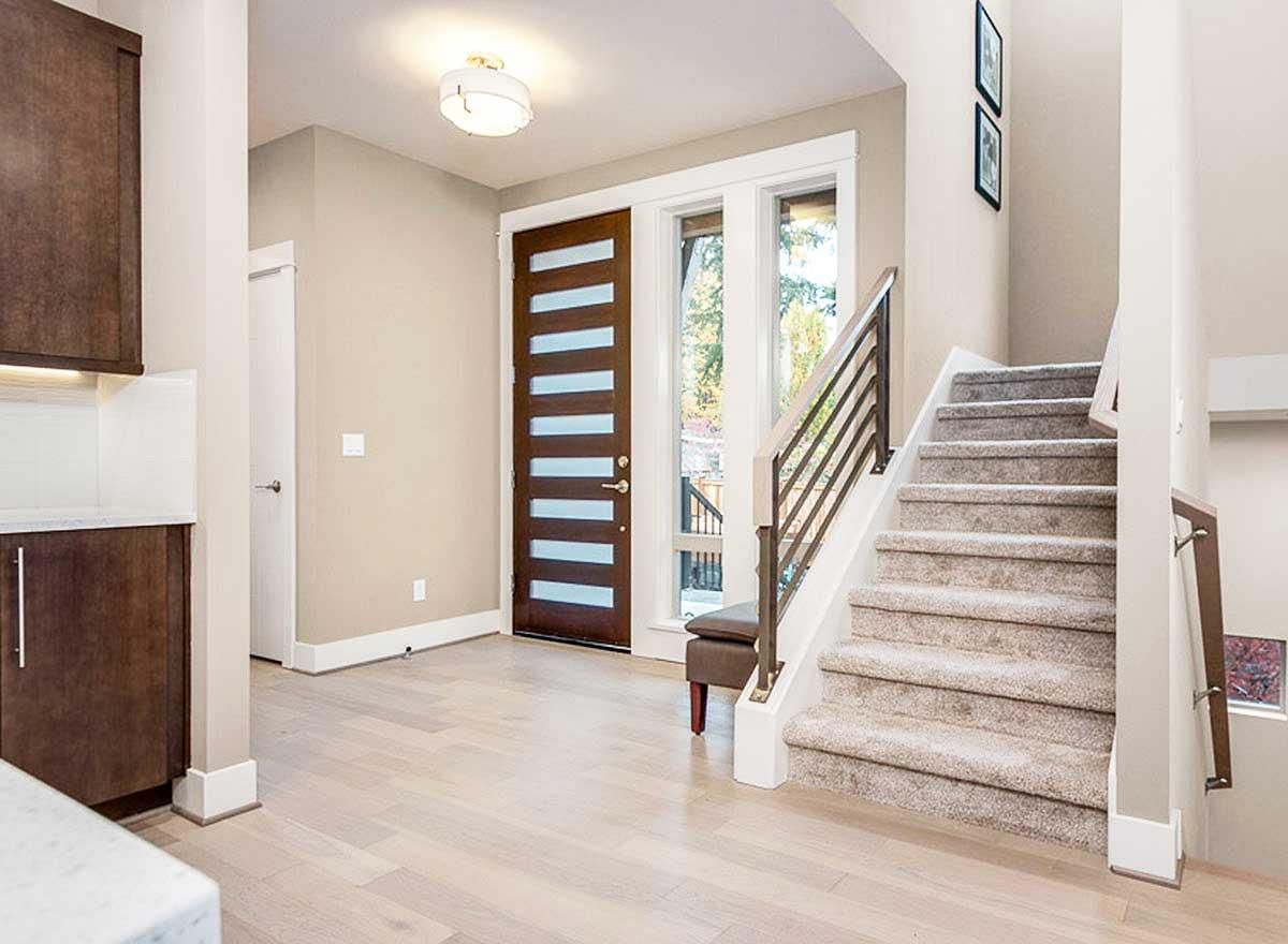 Interior entry hall with modern wood door, large windows, carpeted stairs with metal railing, and dark wood cabinetry.