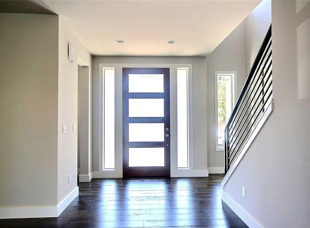 Interior view of a modern entryway featuring a dark wood door with four glass panes, flanked by tall windows, and a staircase with black railings.
