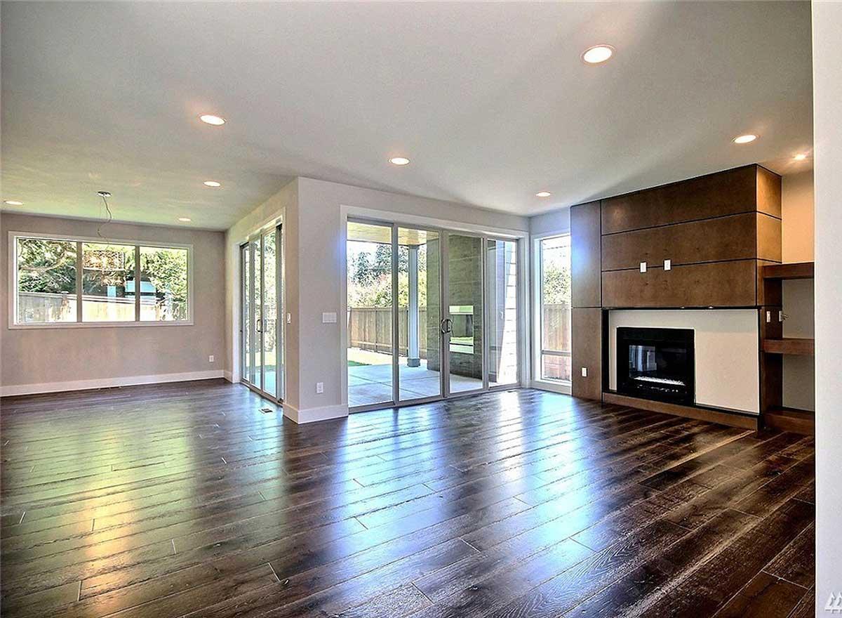 Interior view of a living room with a fireplace, large windows, sliding glass doors to an outdoor patio, and dark wood flooring.