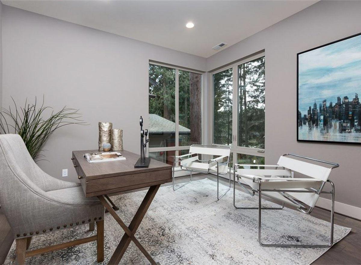 Home office with corner windows, a desk, two chairs, and a cityscape painting on the wall.