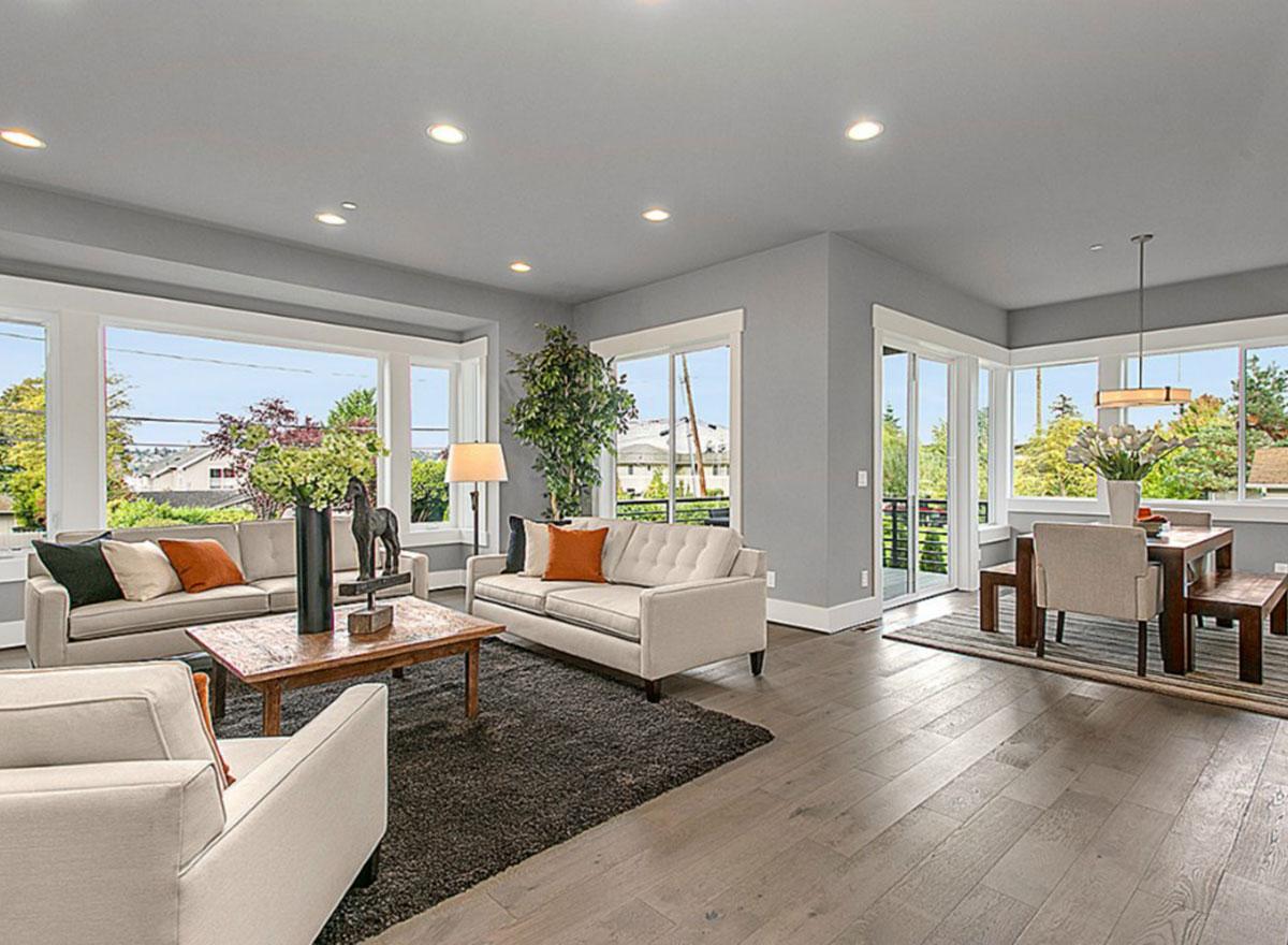 Living and dining area with large windows, neutral sofas, a dark rug, and a wooden dining table with chairs.