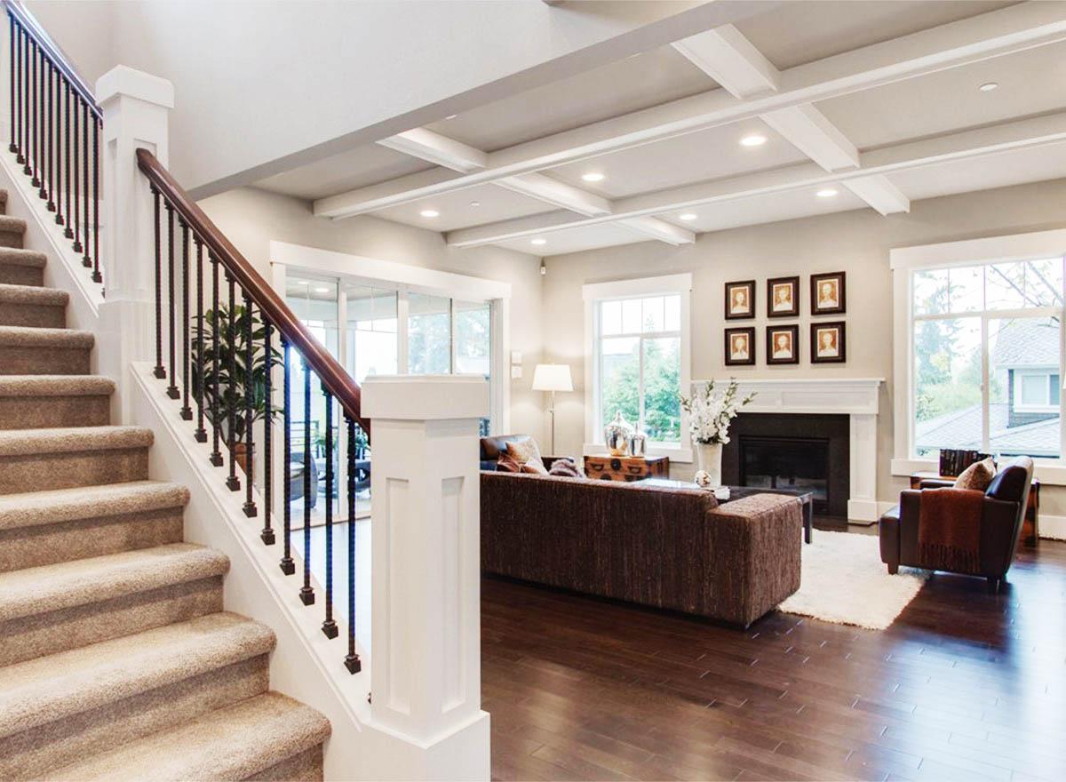 Interior view of a living room featuring a coffered ceiling, fireplace, large windows, and carpeted staircase with ornate railing.