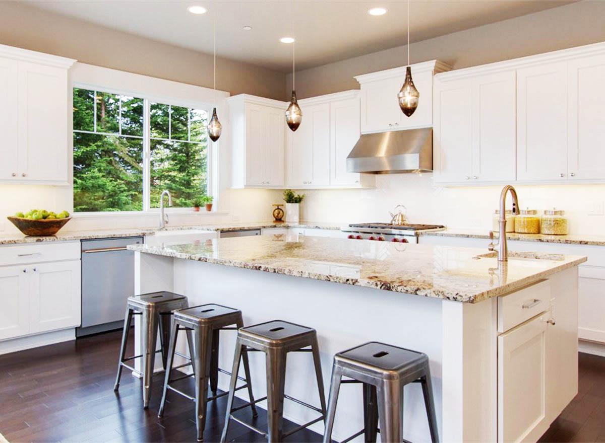 Modern kitchen with white cabinets, granite island with seating, stainless steel appliances, and a large window overlooking trees.