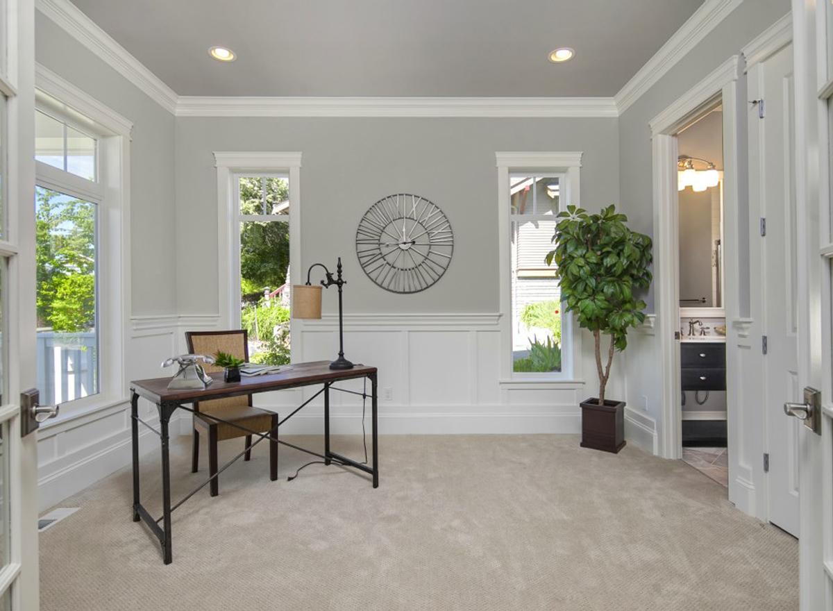 Home office with desk, chair, decorative clock, and large windows overlooking greenery. Doors open to bathroom.