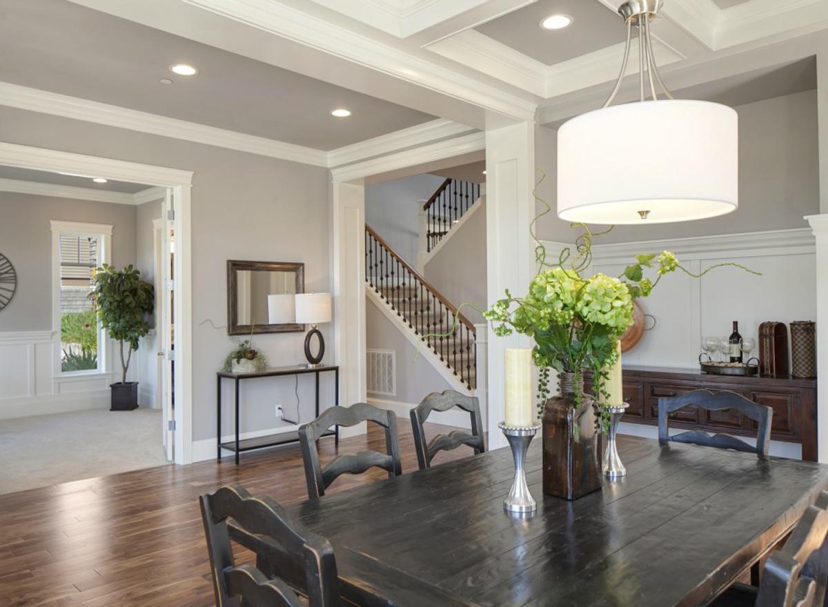 Interior view of dining room with coffered ceiling, dark wood table, staircase with dark spindles, and large window view to living area.