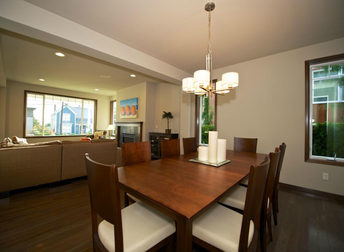 Interior view of a dining room with a large wooden table, upholstered chairs, and a chandelier, adjacent to a living area with a sectional sofa.